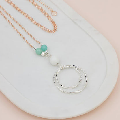 Long silver chain necklace with turquoise and white beads above a pendant of two wavy silver rings